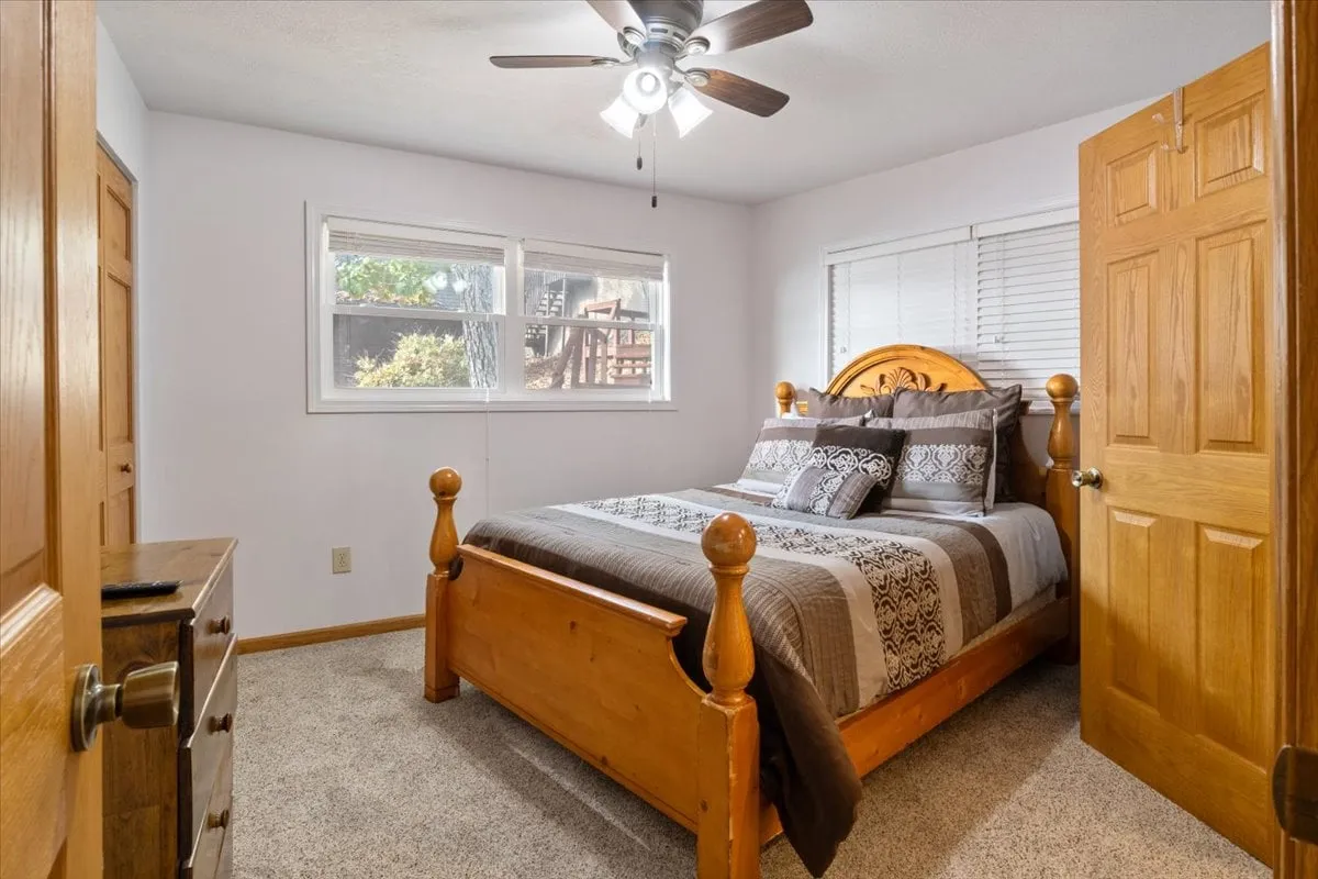 A calming bedroom with a wooden bed frame, patterned bedding, a ceiling fan overhead, and windows that flood the space with natural light. The scene exudes warmth and comfort, ideal for restful sleep and lazy mornings.