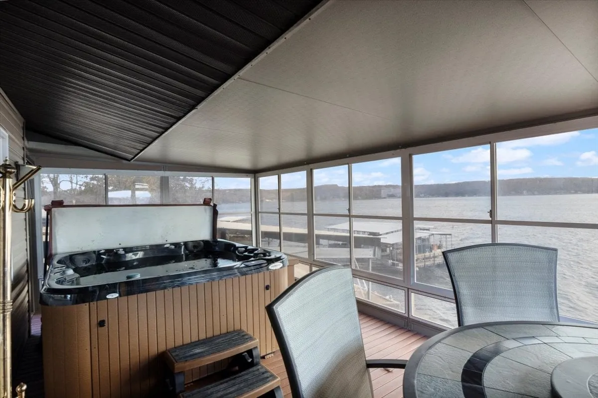 The photo features an airy interior with a hot tub, a dining area, and wide windows framing the lake beyond. The hot tub is prominent in the foreground, while the table invites guests to gather, dine, or enjoy morning coffee with waterfront scenery as the backdrop. The tranquil mood is underscored by the natural lighting and open sightlines to the water.