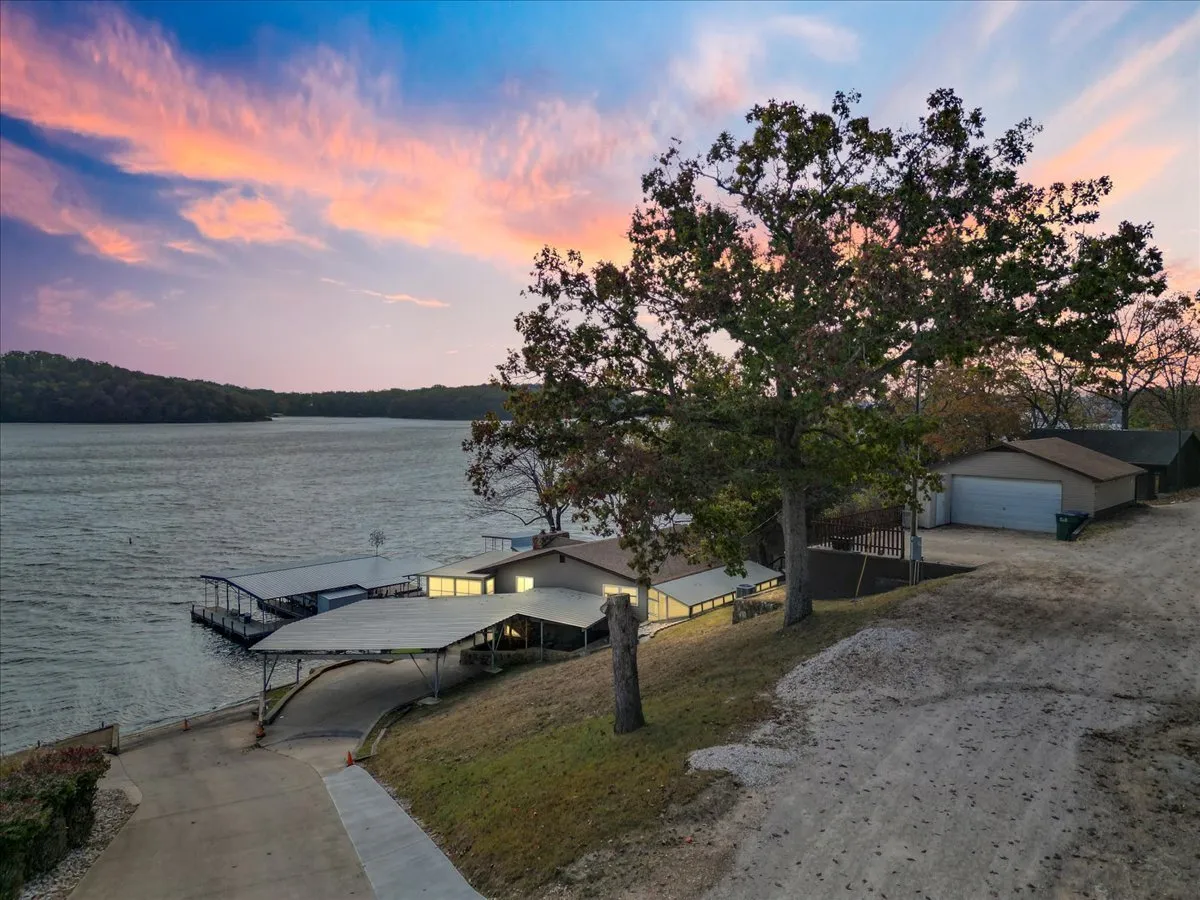 This exterior shot features the lakefront property at sunset, with a large tree providing shade near the docks. The gentle glow of the sunset reflects off the calm water, and the docks are clearly visible, inviting guests to imagine evenings fishing, boating, or relaxing by the water's edge.