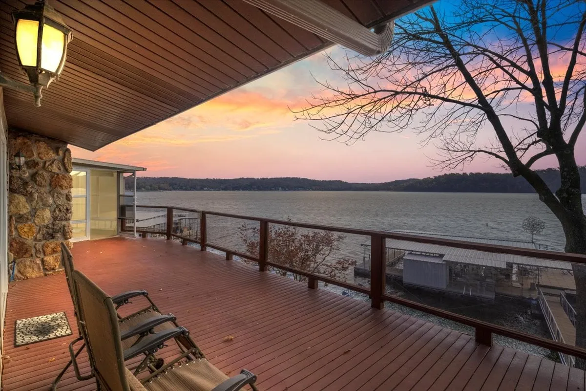 The image showcases a spacious deck overlooking the tranquil waters of Lake of the Ozarks at sunset. Tree branches frame the view, and the vibrant sky casts a warm glow over the lake, inviting moments of relaxation. The scene hints at comfortable seating areas perfect for gathering, and the serene setting evokes the promise of peaceful mornings and magical evenings by the water.