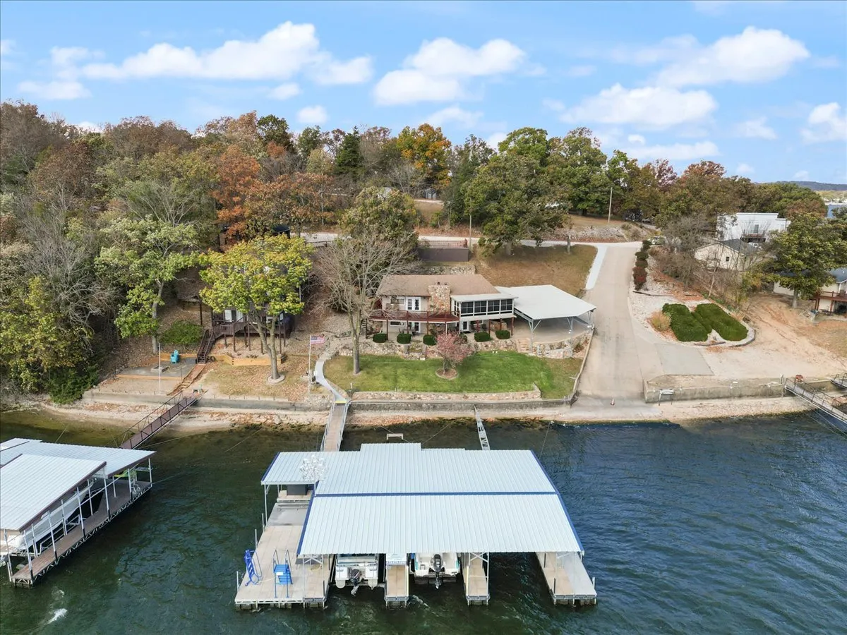 A scenic exterior view of a lakefront property with a private dock stretching out over the water. The dock features multiple boat and PWC slips, and boats are visible moored alongside. Lush greenery surrounds the shoreline, enhancing the inviting atmosphere for outdoor activities.