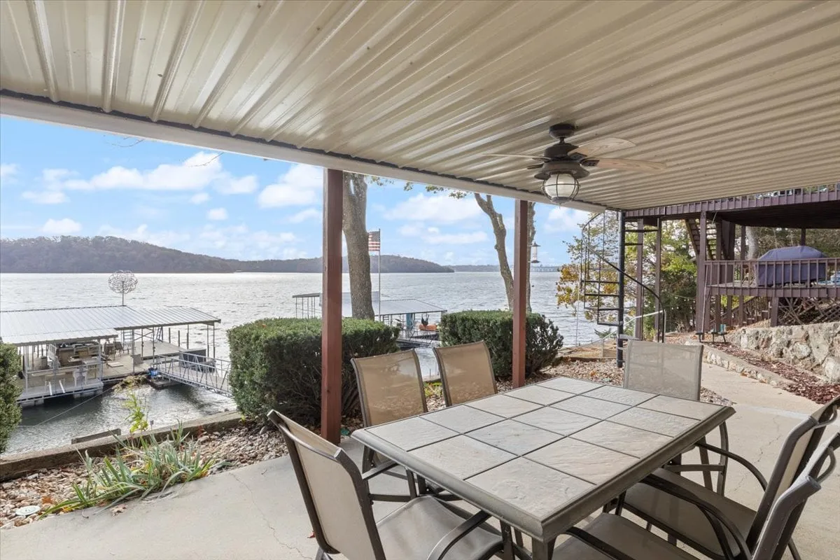 A peaceful exterior shot showcasing an outdoor dining table nestled by the lake, surrounded by greenery and with boathouses in the distance. The image evokes the experience of dining or relaxing outside, overlooking the water, and being at the heart of lakeside community life.