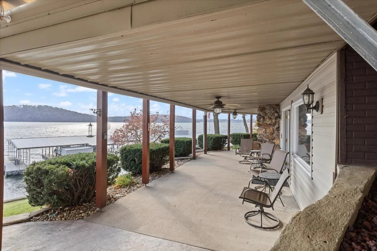 A tranquil patio area with comfortable seating overlooks the lake, surrounded by lush landscaping and a stone wall. The view extends across the calm water, inviting relaxation and connection to nature.