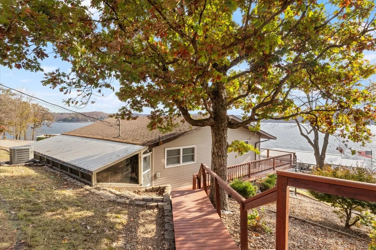 This exterior image features a large tree shading a wooden pathway, the house in view, and a serene expanse of water beyond. The scene captures the transition from the property to the natural surroundings, inviting guests to explore both the house and the outdoors, with space for gatherings and activities like fishing or boating.
