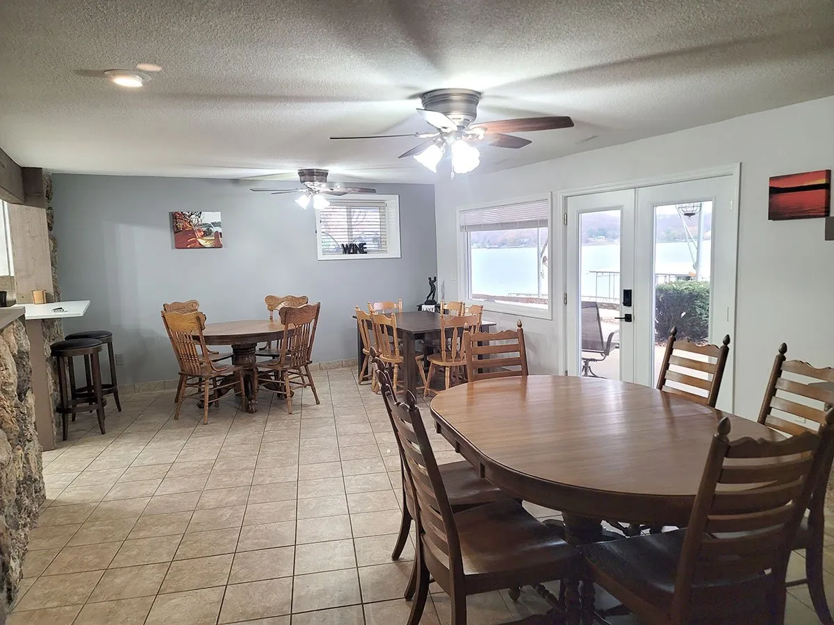 A warm and welcoming dining area with multiple tables set next to expansive windows, allowing natural light to flood the room. Decorative wall art and a bar area add character, and the open-concept layout encourages gatherings for meals, games, and celebrations. The atmosphere is lively, inviting, and perfect for group interaction.
